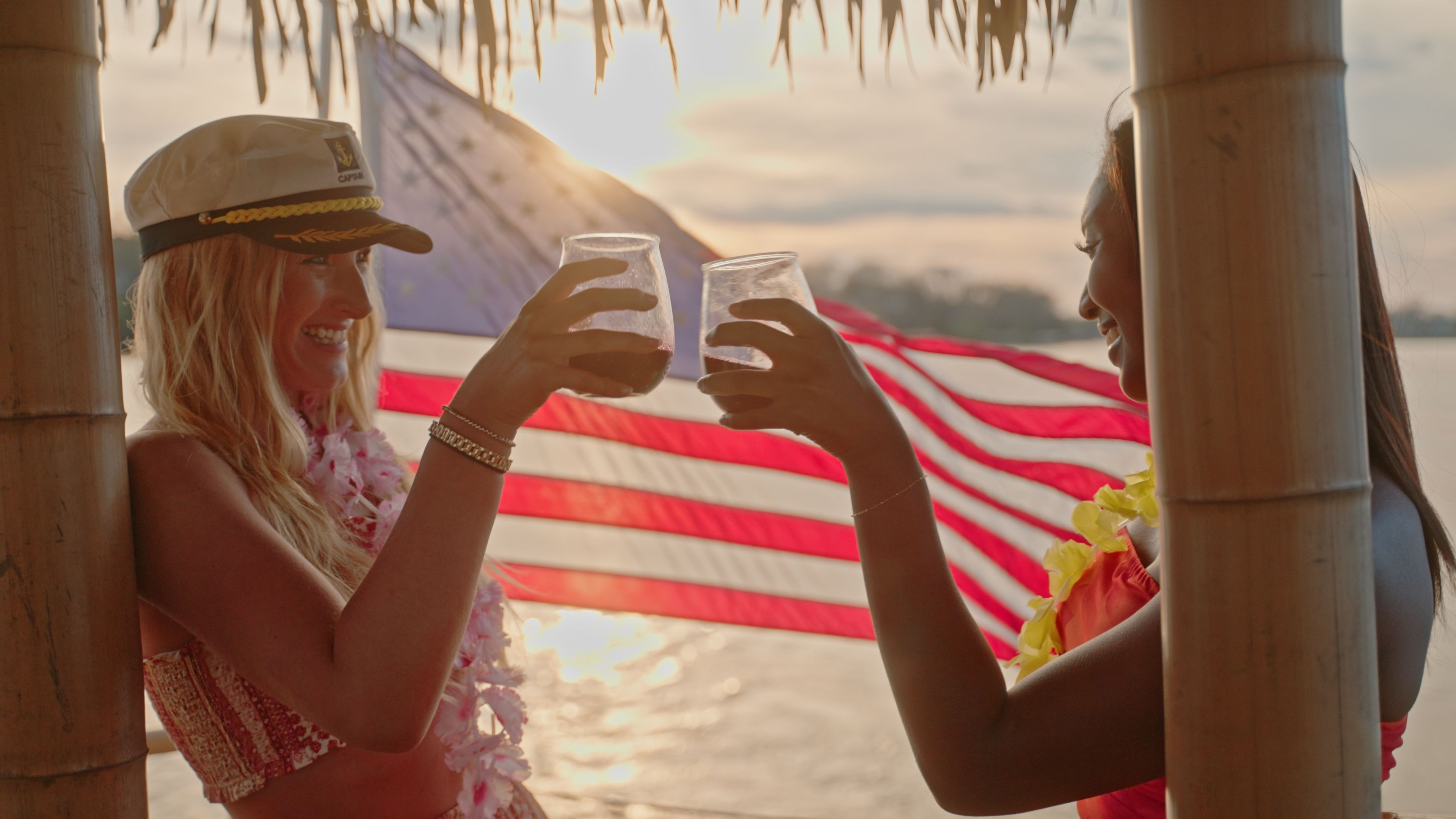 Two people clinking cocktails under a tiki hut at a tropical beach sunset, wearing leis with an American flag waving in the background