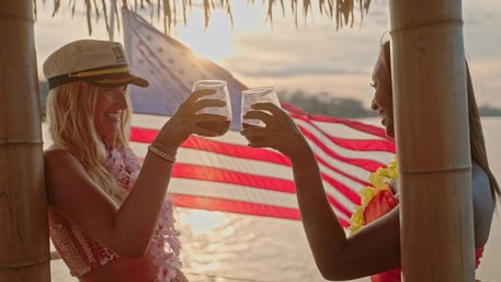 Two people clinking cocktails under a tiki hut at a tropical beach sunset, wearing leis with an American flag waving in the background