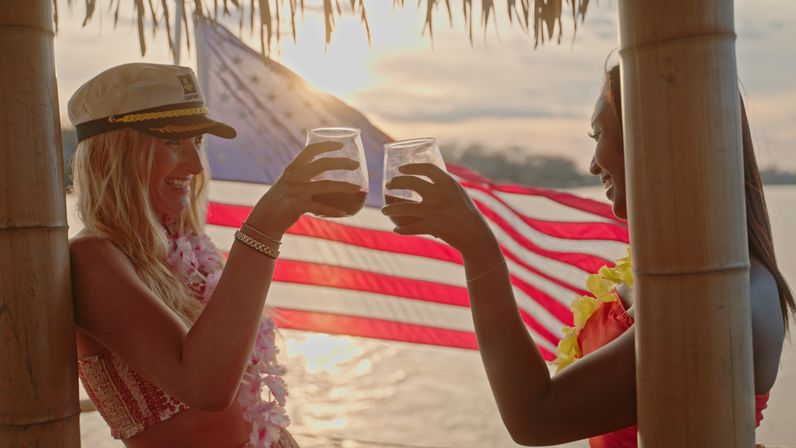 Two people clinking cocktails under a tiki hut at a tropical beach sunset, wearing leis with an American flag waving in the background