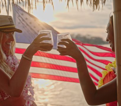 Two people clinking cocktails under a tiki hut at a tropical beach sunset, wearing leis with an American flag waving in the background