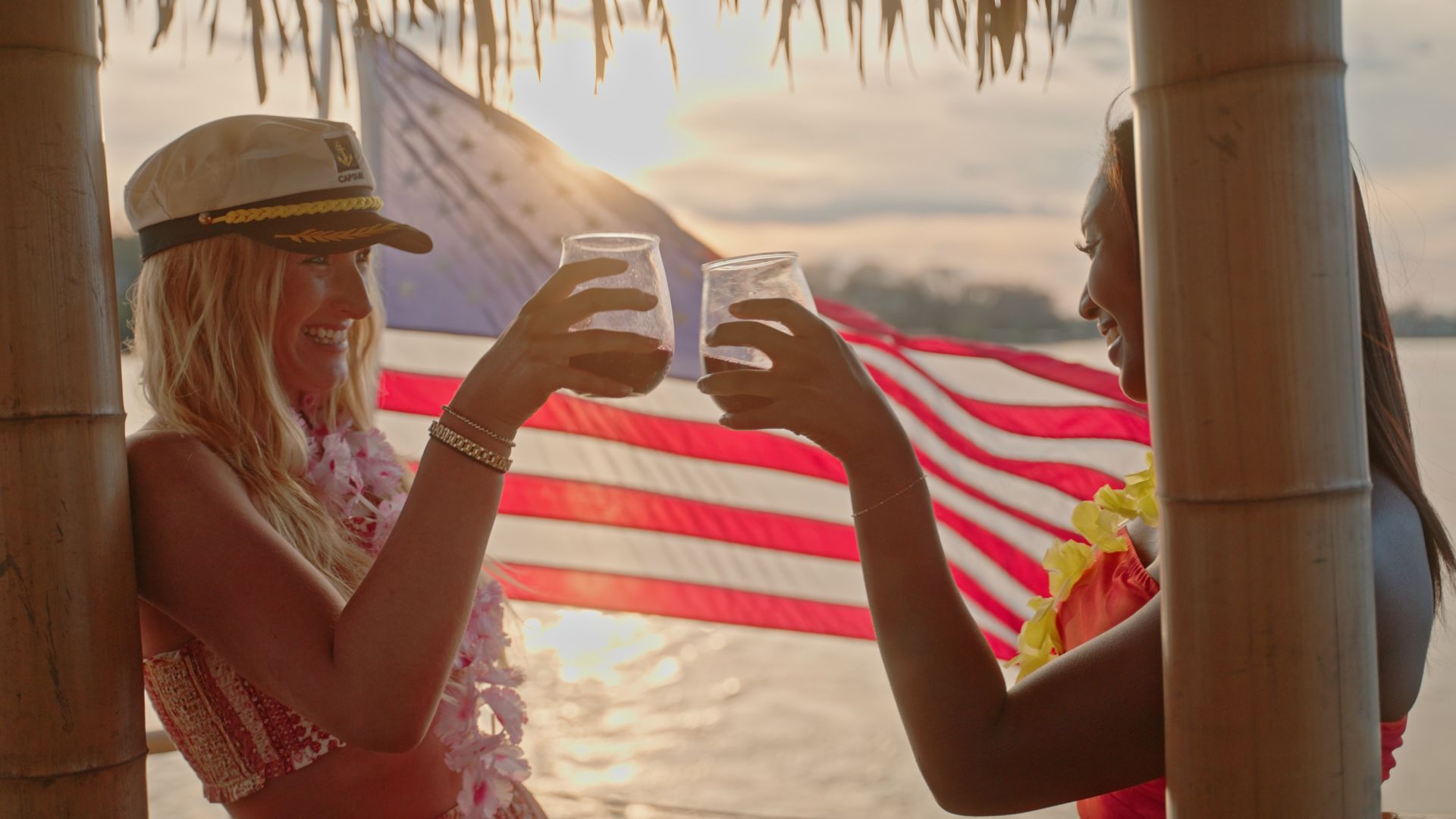 Two people clinking cocktails under a tiki hut at a tropical beach sunset, wearing leis with an American flag waving in the background