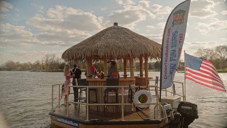 Thatched-roof tiki bar boat floating on a lake with people socializing under the palapa, a waving American flag, tall feather banner, life ring and outboard motor against a cloudy sky.