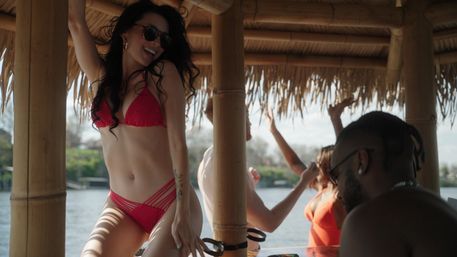 Friends enjoying a tropical boat party under a bamboo tiki hut — woman in a red bikini dancing with sunglasses, others relaxing by the waterfront
