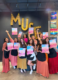 Cheerful group of women at a painting party in a studio, posing under large illuminated marquee letters while holding bright pink-and-blue sunset canvases