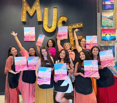 Cheerful group of women at a painting party in a studio, posing under large illuminated marquee letters while holding bright pink-and-blue sunset canvases