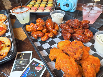 Colorful spread on a wooden table: trays of saucy chicken wings (buffalo and BBQ), ranch dipping sauce, mini flatbreads, rainbow macarons and two cocktails in a casual restaurant setting.