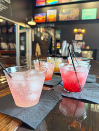 Colorful fizzy cocktails in clear plastic cups with ice and black straws lined on black napkins atop a wooden bar counter, with a blurred cozy bar interior and colorful wall art in the background.