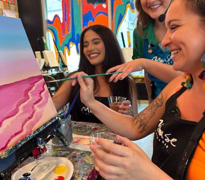 Three smiling people at a paint-and-sip art studio painting a pink coastal canvas, holding brushes and drinks in a colorful, mural-filled interior.