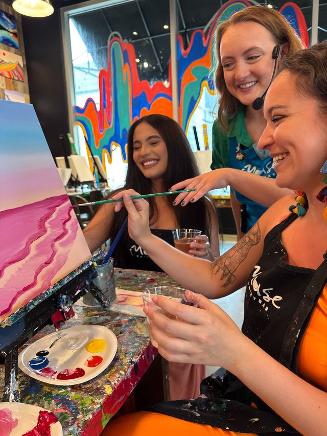 Three smiling people at a paint-and-sip art studio painting a pink coastal canvas, holding brushes and drinks in a colorful, mural-filled interior.