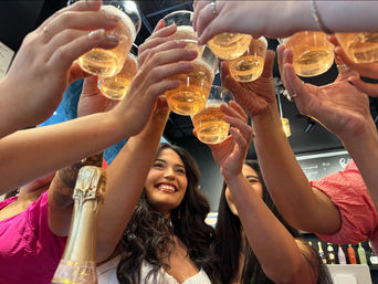 Group of friends toasting with clear champagne cups over a smiling woman in a lively indoor bar setting, bubbles and a sparkling wine bottle visible.