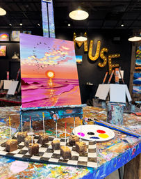 Vibrant sunset beach painting on an easel in a colorful, paint‑splattered art studio, with a palette and brushes nearby and a checkered tray of brownie bites on toothpicks in the foreground — playful painting workshop vibe.