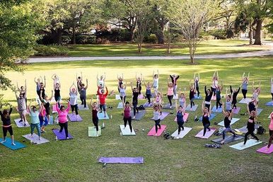 Large outdoor yoga class in a sunny park — dozens practicing standing poses on colorful mats across a grassy lawn surrounded by trees