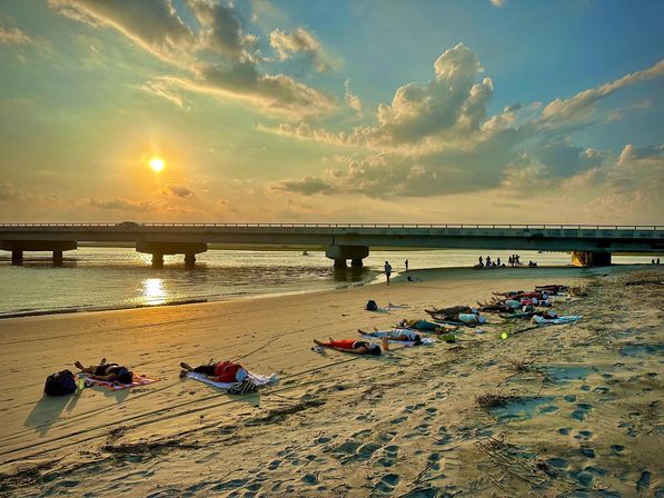 Sunset over a sandy beach with people lying on towels near a low concrete bridge spanning calm water, golden sun and dramatic clouds reflecting on the shoreline.