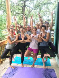 Cheerful group of women doing tree pose on colorful yoga mats on a shaded wooden deck under large oak trees — outdoor group yoga session