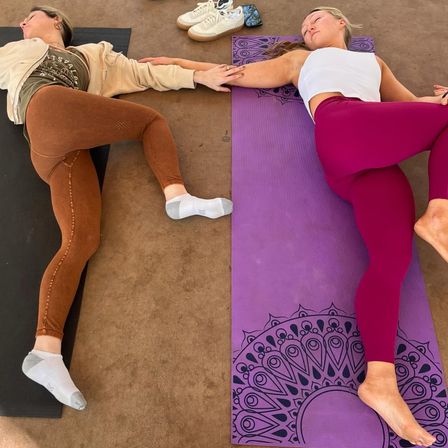 Two people doing a partner yoga supine twist on purple mandala and black mats on a sunlit patio, holding hands with sneakers and a phone nearby