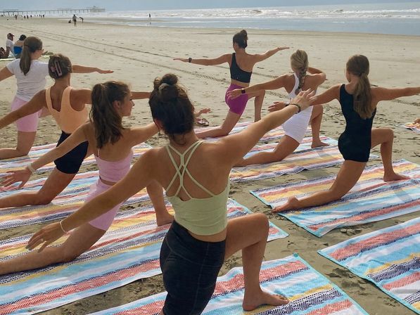 Group of women doing beach yoga on colorful striped towels along a sandy shoreline, with ocean waves and a distant pier in the background
