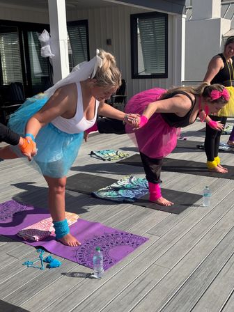 Group of women in bright tutus and leg warmers practicing partnered balancing yoga on a sunny outdoor deck — one wearing a white veil, colorful mats and water bottles nearby.
