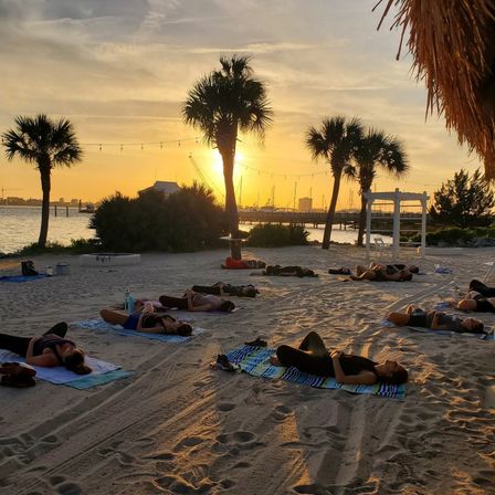 Sunset beach yoga session with people resting on towels on a palm-lined sandy shore, string lights overhead and a marina silhouetted against a golden harbor sky.