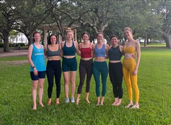 Seven women in colorful activewear smiling and standing arm-in-arm on a green lawn beneath sprawling oak trees with a gazebo in the background.
