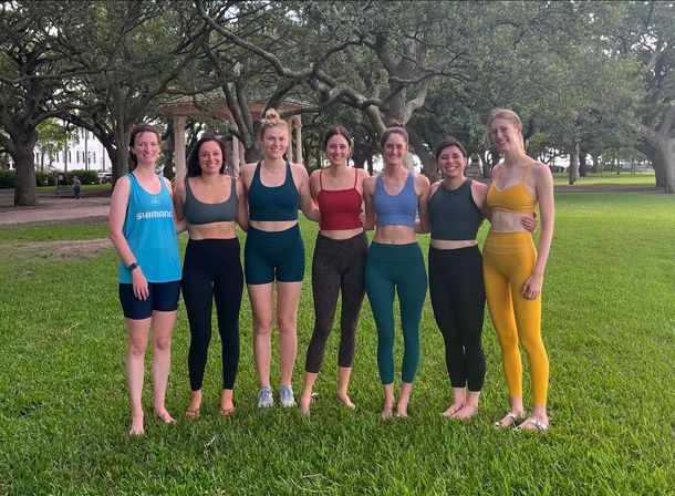 Seven women in colorful activewear smiling and standing arm-in-arm on a green lawn beneath sprawling oak trees with a gazebo in the background.
