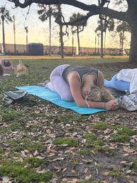 Outdoor park yoga at sunset — person in child's pose on a blue mat under oak branches with palm trees in the background, sneakers and a bag beside the mat, group practice vibe.