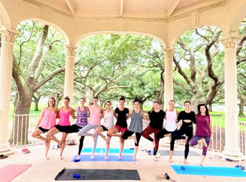 Group outdoor yoga class practicing tree pose on mats inside a park gazebo beneath sprawling live oak trees