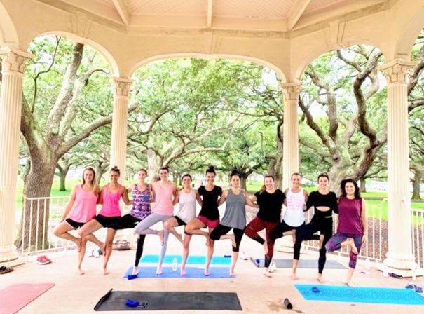 Group outdoor yoga class practicing tree pose on mats inside a park gazebo beneath sprawling live oak trees