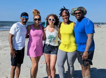 Five adults smiling arm-in-arm on a sunny sandy beach by the ocean, wearing colorful summer outfits and sunglasses — playful group photo on the shore.