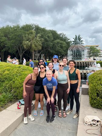 Group of women in athletic wear smiling and posing in front of a pineapple-shaped fountain at a leafy public park under a cloudy sky with palm trees and visitors in the background.