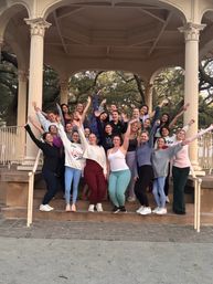 Cheerful group of women in activewear posing with arms raised on the steps of a cream-columned park gazebo bandstand surrounded by live oak trees — outdoor group photo for a fitness or social meetup.