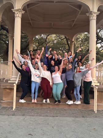 Cheerful group of women in activewear posing with arms raised on the steps of a cream-columned park gazebo bandstand surrounded by live oak trees — outdoor group photo for a fitness or social meetup.