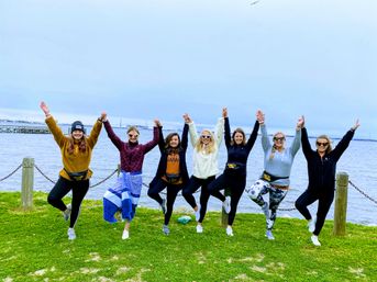 Cheerful group of seven women in activewear holding hands and balancing in tree pose on a grassy waterfront park by a calm bay under an overcast sky.
