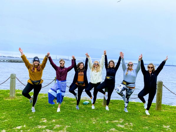 Cheerful group of seven women in activewear holding hands and balancing in tree pose on a grassy waterfront park by a calm bay under an overcast sky.