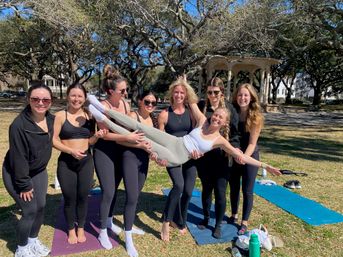 Eight women in activewear smiling in a sunny park, hoisting a friend horizontally above yoga mats near a decorative gazebo and sprawling oak trees — outdoor yoga and fitness meetup.