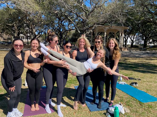 Eight women in activewear smiling in a sunny park, hoisting a friend horizontally above yoga mats near a decorative gazebo and sprawling oak trees — outdoor yoga and fitness meetup.