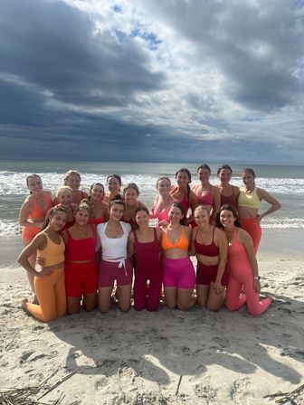 Group of women in bright pink, red and orange activewear posing and smiling on a sandy ocean beach with dramatic cloudy sky overhead