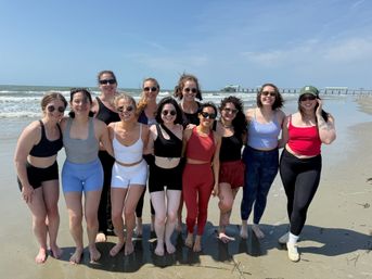 Smiling group of friends in colorful active swimwear and sunglasses posing barefoot on a sunny sandy beach with ocean waves and a pier in the background.