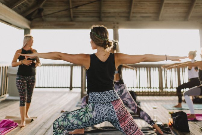 Sunlit outdoor yoga class on a wooden waterfront pavilion, students in colorful leggings holding Warrior II pose with arms extended.