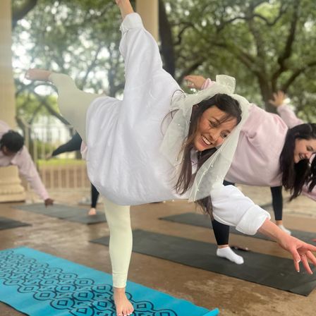 Smiling woman in a sheer bridal veil balances on one leg during a playful outdoor yoga class on a blue patterned mat in a tree-lined park pavilion, friends practicing poses behind her.