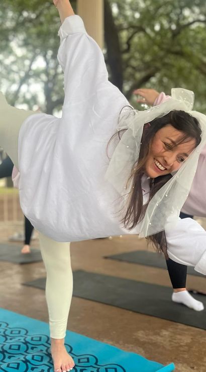 Smiling woman in a sheer bridal veil balances on one leg during a playful outdoor yoga class on a blue patterned mat in a tree-lined park pavilion, friends practicing poses behind her.