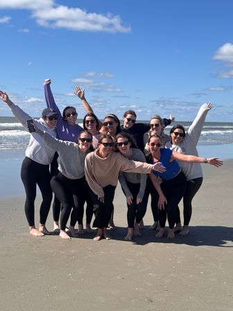 Cheerful group of friends in casual clothes and sunglasses, barefoot on a sandy beach with arms raised, ocean waves and a bright blue sky with scattered clouds in the background.