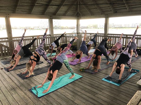 Group outdoor yoga class on a wooden waterfront pavilion, participants on colorful mats practicing three‑legged downward‑dog poses with a coastal pier and water view beyond.
