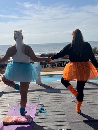 Two people holding hands practicing tree pose in blue and orange tutus and matching leg warmers on a sunlit oceanfront deck by a pool.