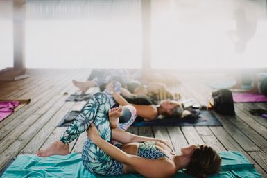 Outdoor yoga class on a sunlit wooden deck in an open-air pavilion — participants in colorful leggings stretching and relaxing on mats.