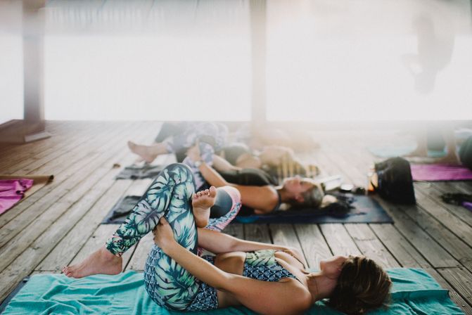 Outdoor yoga class on a sunlit wooden deck in an open-air pavilion — participants in colorful leggings stretching and relaxing on mats.