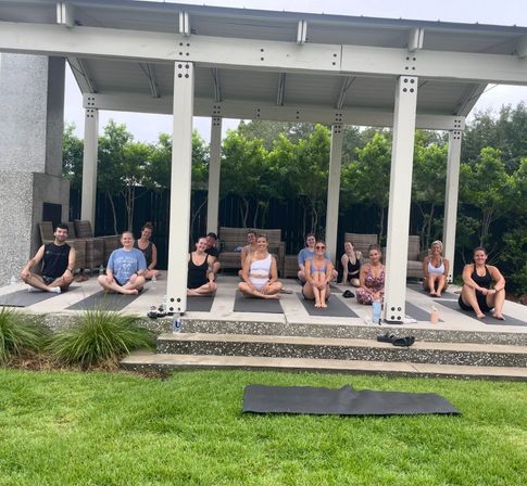 Outdoor group yoga class on mats under a white pavilion in a leafy backyard, participants seated and smiling after a session — relaxed community fitness scene.