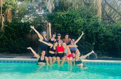 Group of friends in swimwear posing and waving at a sunny backyard pool with palm trees and lush green foliage