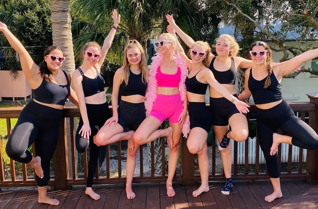 Seven friends on a sunny wooden deck with palm trees, wearing black activewear and heart-shaped sunglasses; center person in bright pink outfit and feather boa strikes a playful pose as the group smiles and lifts a leg in celebration.