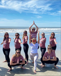 Nine women striking yoga poses on a sunny sandy beach by the ocean, wearing matching mauve shirts and sunglasses with the bride in a white tee at center, gentle waves and blue sky in the background.
