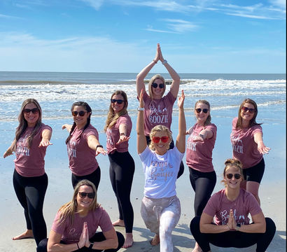Nine women striking yoga poses on a sunny sandy beach by the ocean, wearing matching mauve shirts and sunglasses with the bride in a white tee at center, gentle waves and blue sky in the background.